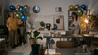 Two diverse women spending morning in office, preparing for colleagues birthday, decorating room with Happy Birthday Banner and balloons