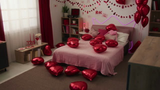 Interior of bedroom prepared for Valentines day showing double bed with pink bedspread decorated with heart-shaped balloons, paper chains and posters on wall, deep red curtains