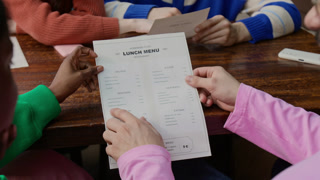 Over the shoulder view of two multiethnic young friends holding menu together and choosing what to order while visiting restaurant with unrecognisable friends sitting in front of them