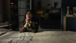 Gen Z woman wearing brown shirt sitting on floor next to bed with hands clasped in front of her chin and lowering head during morning prayer in dimly lit bedroom
