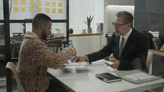 Gen Z man receiving application form from middle-aged male HR specialist and signing it after successfully going through job interview in modern office