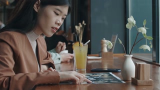 Young Asian woman sitting at table with cocktail in busy restaurant and looking at images of dishes on menu, choosing what else to order