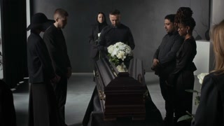 Mourners in black standing gravely with bowed heads around dark wooden coffin adorned with white flowers at funeral