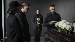Mourners in black standing around coffin and holding white roses, while priest reading from book and conducting funeral service