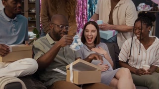 African American man taking gift box with white ribbon from Black female friend, opening it and finding bootees for expected child, his Caucasian pregnant wife sitting next to him during home party