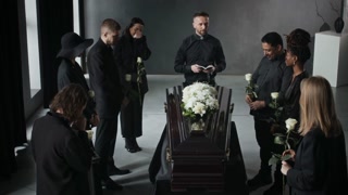 High angle shot of mourners in black, each holding white rose, surrounding flower-draped coffin while priest reading from book, conducting funeral service.