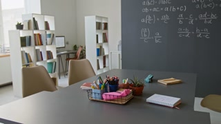 No people shot of interior of well-lit classroom with large gray table with school supplies, and chalkboard with mathematical formulas
