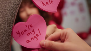 Close-up rack focus shot of cheerful girl with curly hair gently putting pink Valentine post-it note with romantic message on mirror