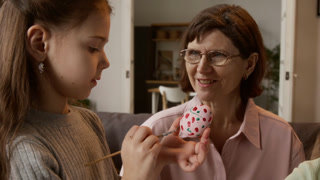 Little Caucasian girl using painting brush while decorating Easter egg with grandmother and little sister in cozy modern living room