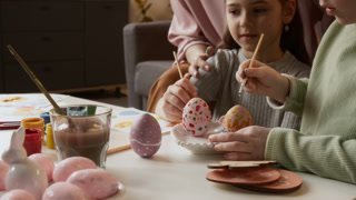Little Caucasian girl sitting at table in living room and painting using brush and gouache on dyed Easter egg while preparing decorations with grandmother and sister at home