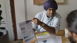 Indian male manager holding sheet of paper and pointing at charts and graphs while explaining business report to colleague at work desk