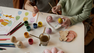 High angle cropped shot of hands of unrecognizable little girls dipping brushes in gouache paint and drawing pattern on dyed eggs while preparing Easter decorations at table