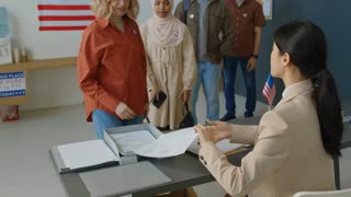 Tilt up shot of young female election inspector sitting at desk and giving voting slips to diverse Americans coming to polling place