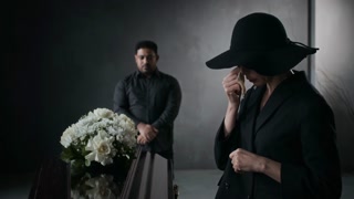 Senior woman in black, wearing hat and wiping tears from her eyes, standing near flower-adorned coffin at funeral