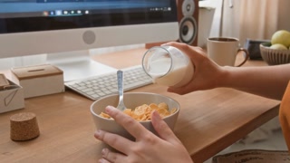Close-up view of hands of unknown girl with disability pouring milk into bowl with cornflakes at computer desk