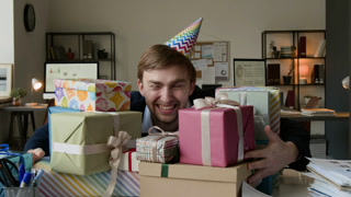 Zoom in portrait of cheerful young man with birthday hat smiling at camera wrapping hands around colourful present boxes placed on his office desk