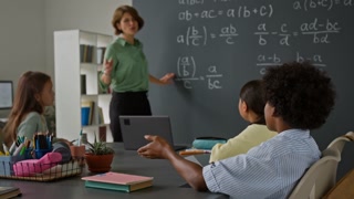 Female teacher pointing to equation on a chalkboard filled with mathematical formulas as student raising hand in class
