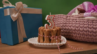 Close-up zoom in view of dog birthday cake representing bone-shaped cookies tied together with string and decorated with numeral candle between blue gift box and pink knitted pet bed on dresser