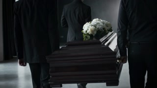 Back view of four unrecognizable men in black suits solemnly carry white-flowered, dark wooden coffin indoors at funeral
