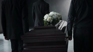 Back view of four unrecognizable men in black suits solemnly carry white-flowered, dark wooden coffin indoors at funeral