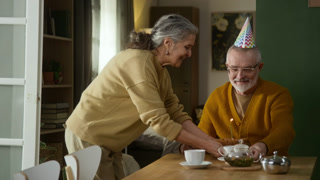 Positive senior lady placing festive cake with burning candle on table in front of surprised husband wearing birthday hat