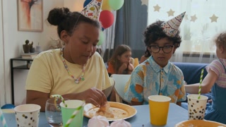 Girl smearing creamy cake on friends face, they smiling and looking at camera while sitting at holiday table together