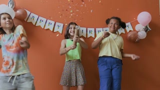 Birthday girl blowing up party popper and enjoying confetti rain with two friends against peach painted wall decorated with balloons