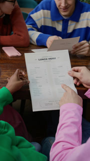 Vertical over the shoulder shot of two multiethnic friends reading lunch menu while visiting cafe with Caucasian couple sitting in front of them