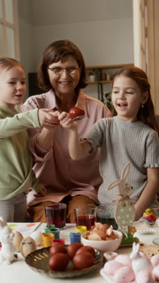 Vertical shot of beautiful little Caucasian sisters and their mature grandmother dying Easter egg in food coloring, enjoying preparing for holiday together at home