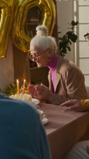 Vertical view of senior man bringing festive cake for stylish elderly woman celebrating 70th birthday with friends at holiday table, she blowing out candles out
