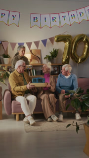 Vertical view of elegant elderly woman celebrating 70th birthday with loving friends who giving her presents and wishing health and happiness in apartment decorated with helium balloons and buntings