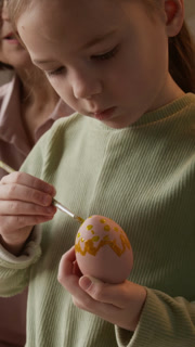 Vertical tilt down shot of little Caucasian girl child applying yellow paint on pink Easter egg using brush with help of her caring mature grandmother in background