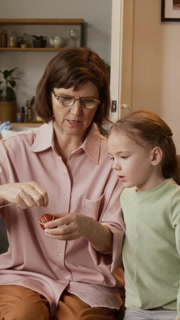 Vertical shot of mature Caucasian grandmother explaining to inspired little granddaughter how to wrap thread around Easter egg, preparing holiday decorations together