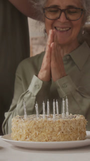 Vertical shot of Caucasian grey-haired senior woman wearing earrings, glasses and green shirt blowing out candles on birthday cake, then clapping hands with happiness during family party