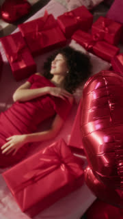 Vertical portrait from above of curly-haired gen Z woman in elegant dress looking at camera lying on her back on bed surrounded by red gift boxes tied with ribbons
