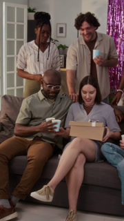 Vertical shot of happy diverse couple getting gift box from young man, woman opening it and taking out knitted blanket while celebrating baby shower with friends