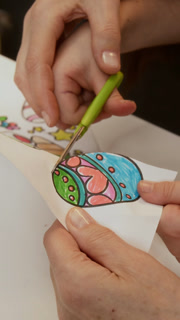 Vertical cropped shot of hands of unrecognizable adult helping little child to cut out colored Easter egg drawing, engaged in creative activity during preparation for spring holiday together