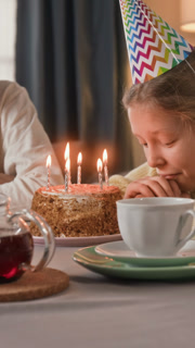 Vertical shot of pensive birthday girl watching candles lit on festive cake with smile on her face while celebrating with unknown brother