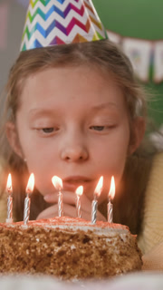 Vertical shot of dreamy child with birthday hat whispering her wish pensively looking at candles lit on sponge cake