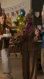 Vertical view of elegant elderly woman using smartphone with two friends standing nearby when her husband bringing cream cake with lit candles during birthday party