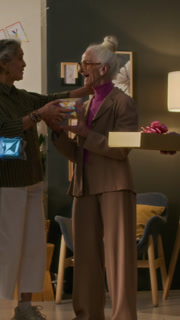 Vertical view of senior friends congratulating cheerful birthday woman on 70th anniversary and giving her gift boxes in living room