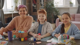 Portrait of happy mother, daughter and son sitting together at table with dyed Easter eggs and craft supplies, looking at camera and smiling