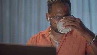 Tilt down shot of black man in wireless earphones using laptop and drinking tea while working remotely from home in evening