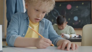 Little kid with blond curly hair putting marks on paper sheet with pencil while doing craft project on lesson in elementary school