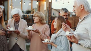 Cheerful mixed-age family members eating birthday cake, smiling and chatting while having dinner party outdoors in backyard decorated with lights