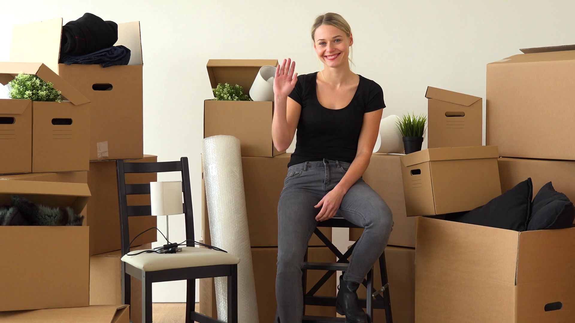 A moving woman sits on a chair in an empty apartment and waves at the camera with a smile ...