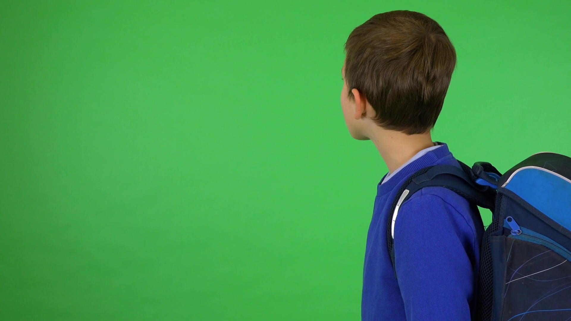 A young cute boy with a schoolbag looks around green screen studio