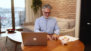 Mature man counting coins, managing personal finance and savings with a laptop and piggy bank for retirement planning at home