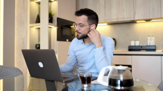 Professional man using wireless earbuds for a video conference call while working remotely from his modern home kitchen office