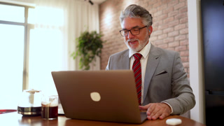 Senior businessman having an online video call meeting on a laptop with earphones from his modern home office with a coffee pot